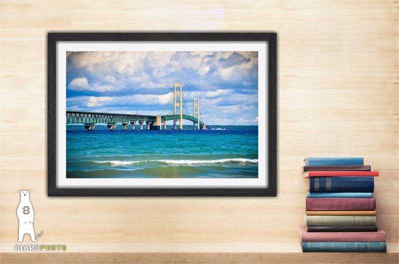May include: A framed photograph of the Mackinac Bridge in Michigan. The bridge is a suspension bridge with a green roadway and yellow towers. The bridge is over a body of water with blue water and white waves. The sky is blue with white clouds.
