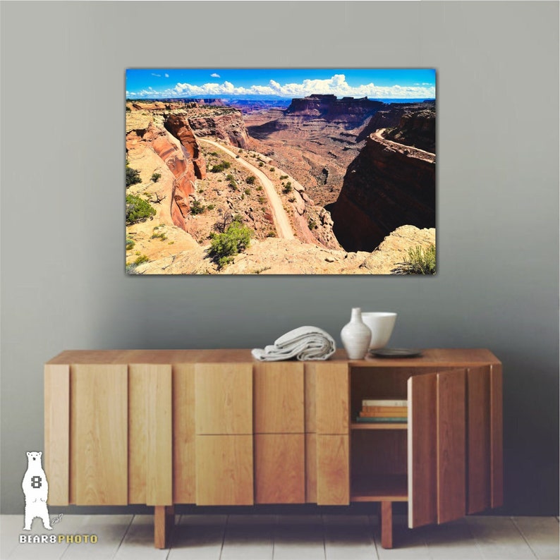 May include: A scenic photograph of a winding dirt road through a canyon in a desert landscape. The road leads to a cliff edge with a view of the canyon below. The sky is blue with white clouds.