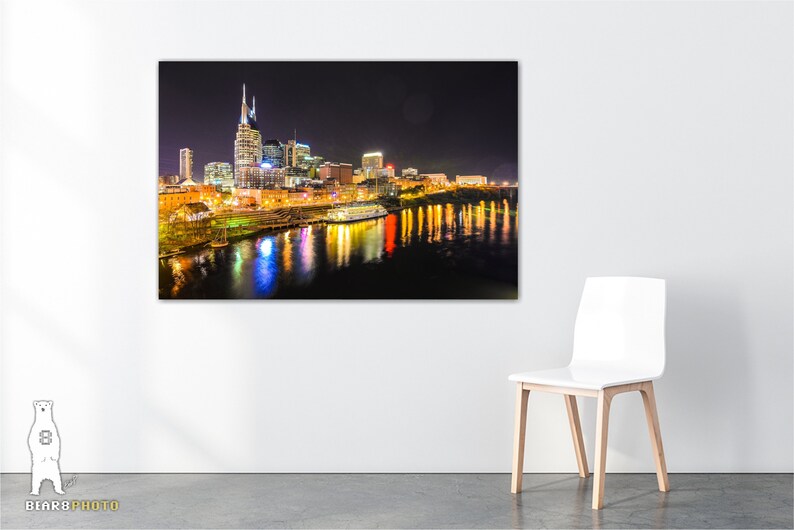 Może przedstawiać: Nocny widok panoramy Nashville z rzeką Cumberland na pierwszym planie. Światła miasta odbijają się w wodzie, tworząc piękny widok.