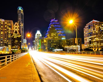 May include: A night time view of a city skyline with tall buildings lit up. The street below is illuminated by car headlights creating streaks of light.