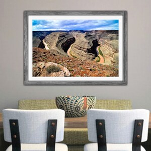 May include: An aerial view of a winding river cutting through a canyon in a desert landscape. The river is a light brown color and the canyon walls are a mix of brown, red, and gray. The sky is blue with white clouds.