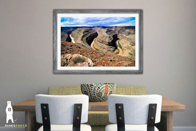 May include: An aerial view of a winding river cutting through a canyon in a desert landscape. The river is a light brown color and the canyon walls are a mix of brown, red, and gray. The sky is blue with white clouds.