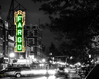 May include: A black and white photograph of a building with a green neon sign that reads "FARGO". The sign is illuminated and there are cars parked in front of the building.