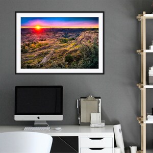 May include: A framed photograph of a sunset over a canyon landscape. The sky is a vibrant mix of pink, orange, and purple, with the sun setting in the distance. The canyon is a rugged, rocky terrain with green vegetation.