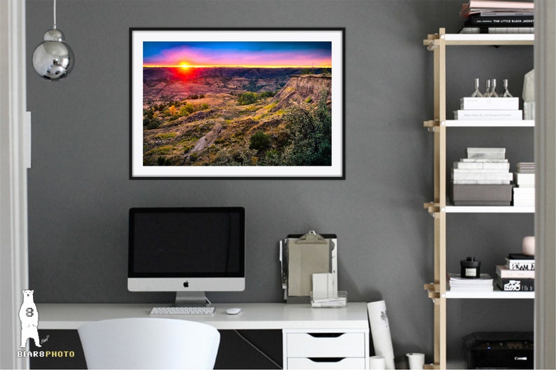 May include: A framed photograph of a sunset over a canyon landscape. The sky is a vibrant mix of pink, orange, and purple, with the sun setting in the distance. The canyon is a rugged, rocky terrain with green vegetation.