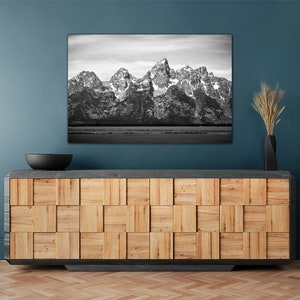 May include: A black and white photograph of a mountain range with snow-capped peaks. The photo is hanging on a teal wall above a wooden cabinet with a black bowl on top.
