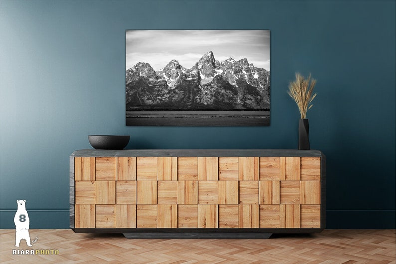 May include: A black and white photograph of a mountain range with snow-capped peaks. The photo is hanging on a teal wall above a wooden cabinet with a black bowl on top.