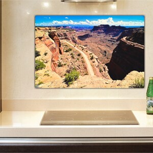 May include: A panoramic view of a winding dirt road through a canyon in a desert landscape. The road is surrounded by red rock cliffs and a blue sky with white clouds.