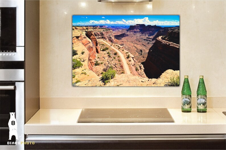 May include: A panoramic view of a winding dirt road through a canyon in a desert landscape. The road is surrounded by red rock cliffs and a blue sky with white clouds.