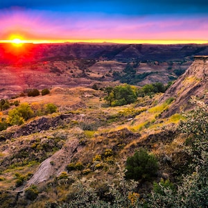 May include: A panoramic view of a rugged landscape with a vibrant sunset in the background. The sun is setting behind a series of hills and valleys, casting a warm glow over the scene. The sky is a mix of pink, orange, and purple hues, creating a dramatic and colorful backdrop.