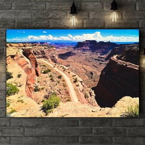 May include: A winding dirt road leads through a canyon with red rock cliffs and a blue sky with white clouds.