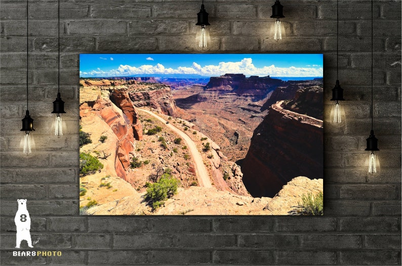 May include: A winding dirt road leads through a canyon with red rock cliffs and a blue sky with white clouds.