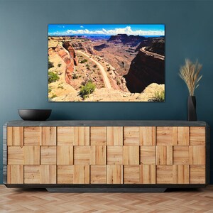 May include: A panoramic photograph of a winding road through a canyon in a desert landscape. The road is surrounded by red rock cliffs and a blue sky with white clouds.