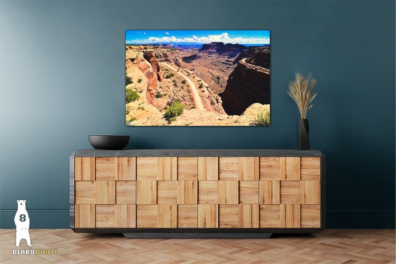 May include: A panoramic photograph of a winding road through a canyon in a desert landscape. The road is surrounded by red rock cliffs and a blue sky with white clouds.