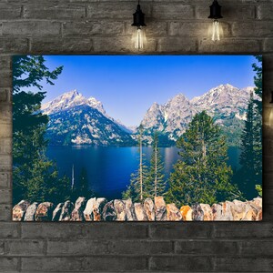 May include: A scenic landscape photograph featuring a mountain range with snow-capped peaks reflected in a still lake. The foreground shows a stone wall and lush green trees.