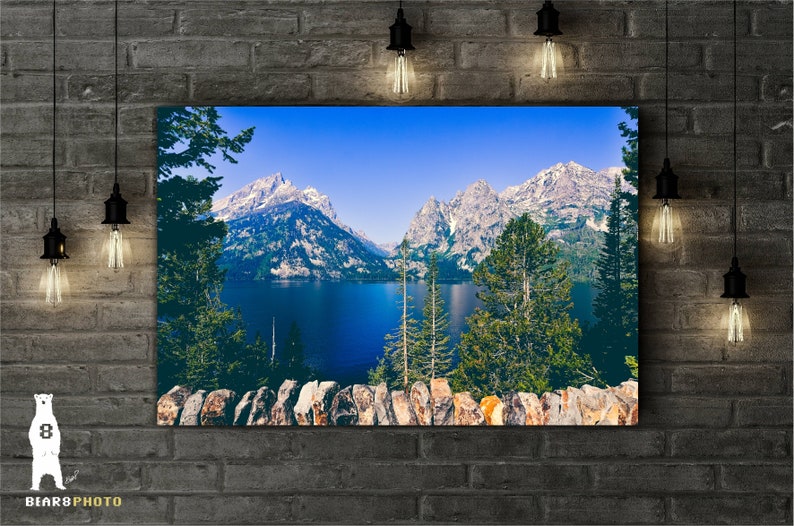 May include: A scenic landscape photograph featuring a mountain range with snow-capped peaks reflected in a still lake. The foreground shows a stone wall and lush green trees.