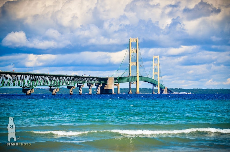 May include: A view of the Mackinac Bridge, a suspension bridge connecting the Upper and Lower Peninsulas of Michigan, with blue water and a cloudy sky in the background.