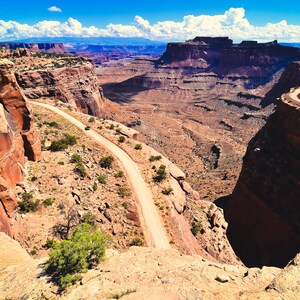 May include: A winding dirt road snakes through a canyon in a desert landscape. The road is surrounded by red rock cliffs and the sky is blue with white clouds.