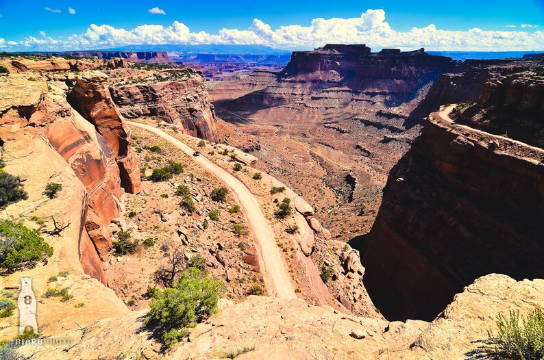 May include: A winding dirt road snakes through a canyon in a desert landscape. The road is surrounded by red rock cliffs and the sky is blue with white clouds.