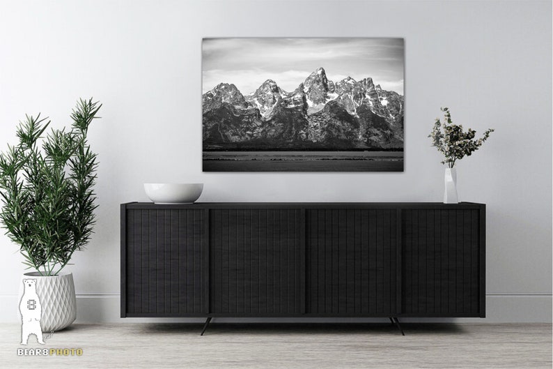 May include: A black and white photograph of a mountain range with snow-capped peaks. The mountains are reflected in a still body of water in the foreground.