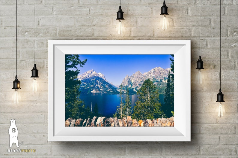 May include: A framed photograph of a scenic mountain lake with snow-capped peaks in the background. The lake is a deep blue color and surrounded by lush green trees. The photo is framed in a white frame.