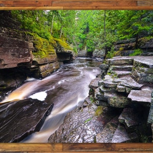 May include: A rocky riverbed with a flowing stream of water. The water is a light brown color and the rocks are a dark gray color. The rocks are covered in moss and lichen. The riverbed is surrounded by lush green trees.