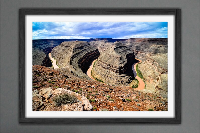 May include: An aerial view of a winding river carving through a canyon in a desert landscape. The river is a light brown color and the canyon walls are a mix of brown, tan, and green.