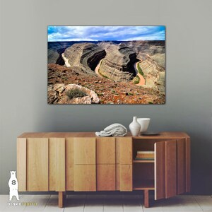 May include: A panoramic view of a winding river snaking through a canyon with layered rock formations. The river is a light brown color and the canyon walls are a mix of brown, tan, and green.