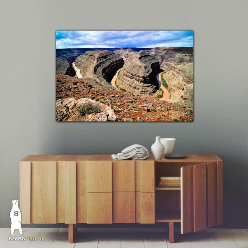 May include: A panoramic view of a winding river snaking through a canyon with layered rock formations. The river is a light brown color and the canyon walls are a mix of brown, tan, and green.