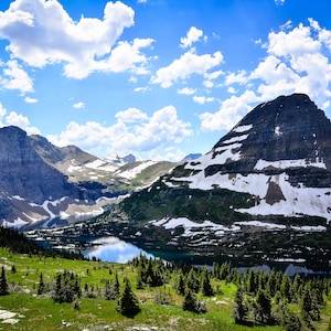 May include: A panoramic view of a mountain range with a lake in the foreground. The mountains are covered in snow and the sky is blue with white clouds.