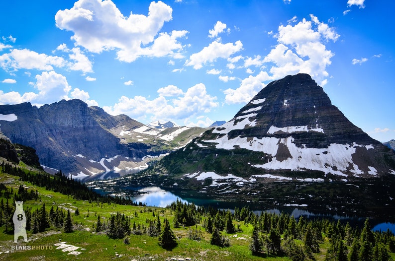 May include: A panoramic view of a mountain range with a lake in the foreground. The mountains are covered in snow and the sky is blue with white clouds.