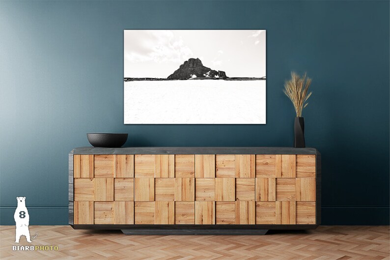 May include: A black and white landscape photograph of a mountain and snow scene. The artwork is displayed above a wooden cabinet with a black bowl and a black vase with dried plants. The cabinet has a geometric design.