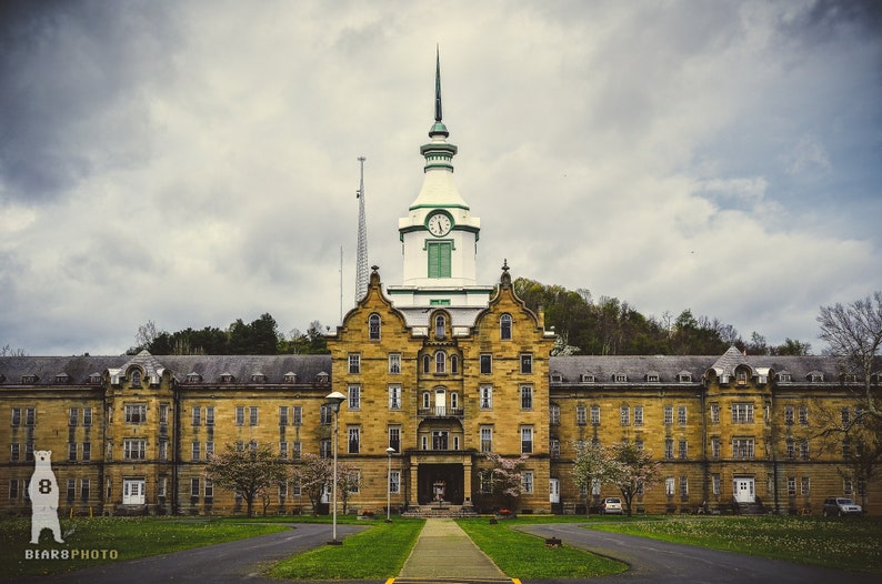 May include: A large, abandoned, yellow brick building with a green and white tower and a clock. The building has many windows and a long, rectangular shape. The building is surrounded by trees and grass.