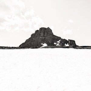 May include: A black and white photograph of a mountain range with snow in the foreground. The mountain is large and rocky, with some snow patches. The sky is overcast.