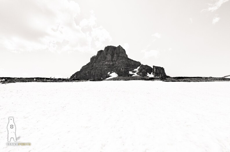 May include: A black and white photograph of a mountain range with snow in the foreground. The mountain is large and rocky, with some snow patches. The sky is overcast.