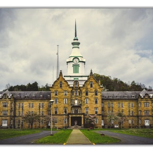 May include: A large, yellow brick building with a clock tower and a green roof. The building has many windows and is surrounded by trees and grass.
