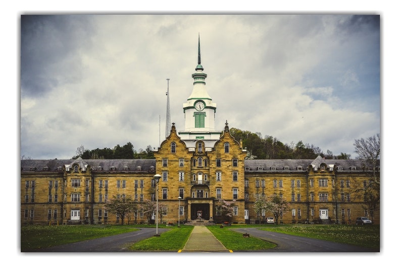 May include: A large, yellow brick building with a clock tower and a green roof. The building has many windows and is surrounded by trees and grass.