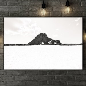 May include: Black and white landscape photograph of a mountain peak with snow at its base. The image is displayed on a white canvas, mounted on a brick wall. Decorative light bulbs hang from the ceiling.