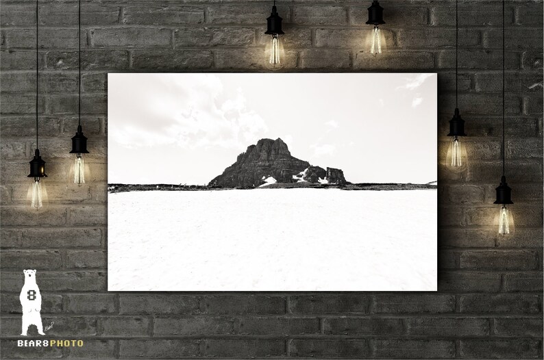 May include: Black and white landscape photograph of a mountain peak with snow at its base. The image is displayed on a white canvas, mounted on a brick wall. Decorative light bulbs hang from the ceiling.