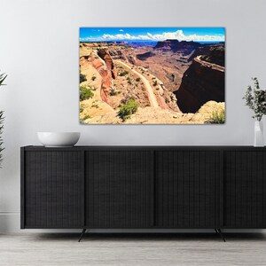May include: A panoramic photograph of a winding road through a canyon in a desert landscape. The road is surrounded by red rock cliffs and the sky is blue with white clouds.