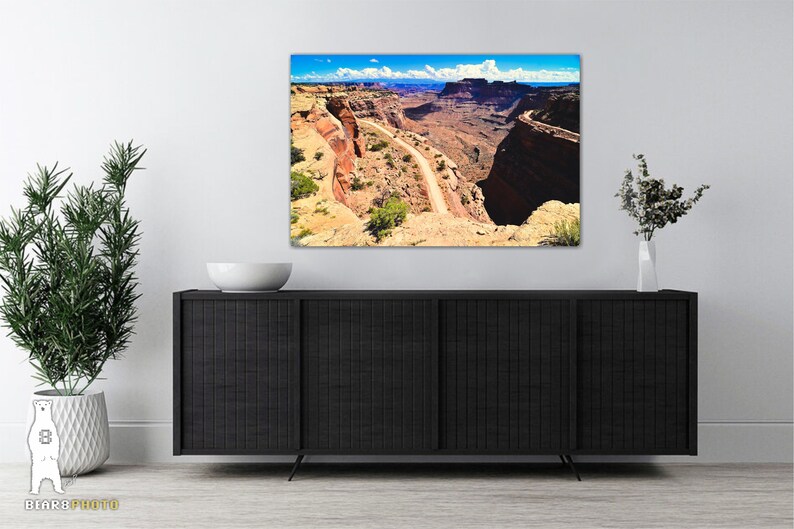 May include: A panoramic photograph of a winding road through a canyon in a desert landscape. The road is surrounded by red rock cliffs and the sky is blue with white clouds.