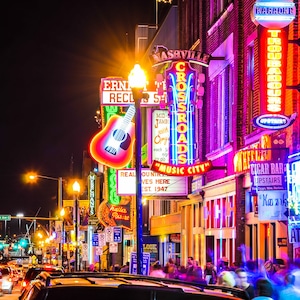 May include: A brightly lit street scene in Nashville, Tennessee at night.  Neon signs with the words "Nashville Crossroads Music City", "Troubadours", "Merchants", "Real Country", "Ernie's Records", and "Cigar Bar" are visible.  The street is lined with buildings and there are people walking on the sidewalk.