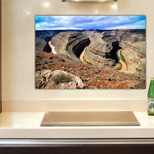 May include: A panoramic photograph of a canyon with a river winding through it. The canyon walls are a mix of brown, tan, and green, and the river is a light brown color. The sky is blue with white clouds.