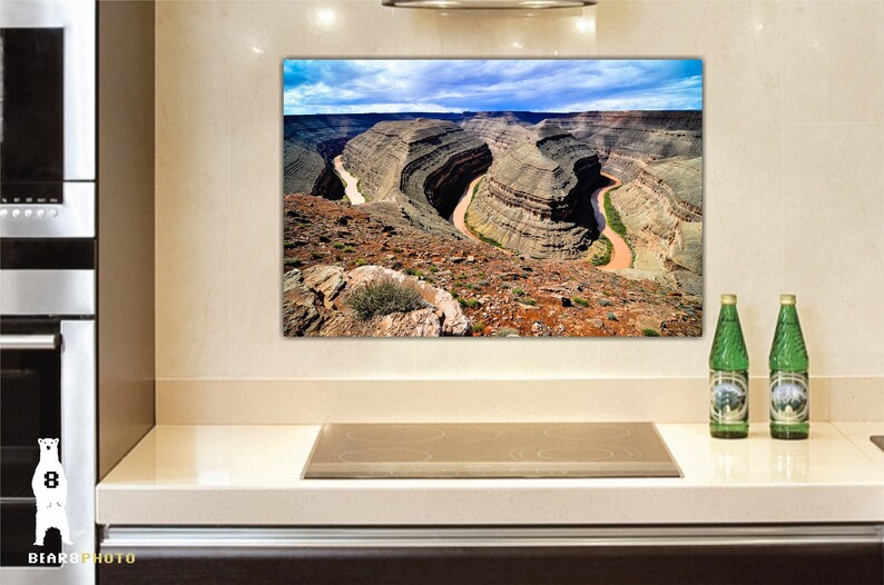 May include: A panoramic photograph of a canyon with a river winding through it. The canyon walls are a mix of brown, tan, and green, and the river is a light brown color. The sky is blue with white clouds.