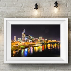 Może przedstawiać: Nocne zdjęcie panoramy Nashville w stanie Tennessee, z rzeką Cumberland w pierwszym planie. Sylwetka miasta jest oświetlona światłami, a rzeka odbija światła. Na rzece widać ł&oacute;dź.