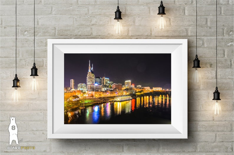 Może przedstawiać: Nocne zdjęcie panoramy Nashville w stanie Tennessee, z rzeką Cumberland w pierwszym planie. Sylwetka miasta jest oświetlona światłami, a rzeka odbija światła. Na rzece widać ł&oacute;dź.