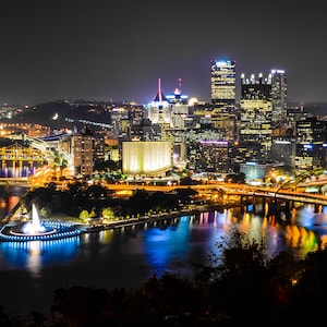 May include: A night view of a city skyline with multiple bridges and a fountain in the foreground. The city is lit up with lights, and the water reflects the lights.