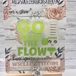 May include: A green glitter badge holder with the text "Go with the flow" and a cartoon oxygen tank with a smiley face. The badge holder is attached to a clear retractable badge reel with a silver clip. The background is a floral design with a wooden plank texture.