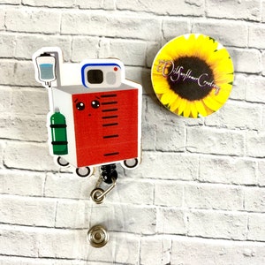 May include: A red and white cartoon medical cart with a green oxygen tank, a clear IV bag, and a black retractable badge reel. The cart has a cute face with eyes and a mouth. The badge reel is attached to a clear plastic badge holder. The background is a white brick wall. The image also includes a sunflower with the text "Wildflower Creations".