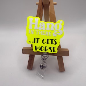 May include: A neon yellow badge reel with the text "Hang in there...IT GETS WORSE" in white lettering. The badge reel is attached to a clear plastic holder and is displayed on a small wooden easel.
